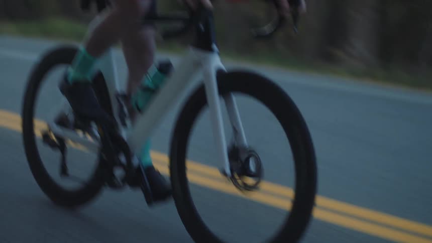 A cyclist in an orange and black long sleeve shirt black shorts and a white helmet is riding a white and black bicycle on a paved road in a wooded area
