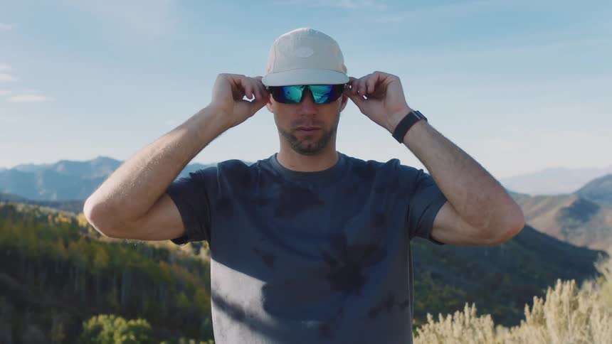 Portrait of a man with sports glasses adjusting his white cap while standing in a natural setting with a mountain valley in the background