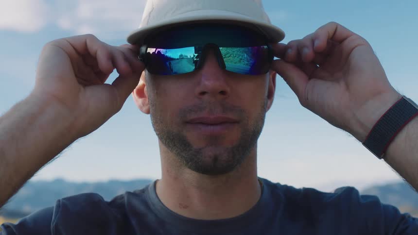 A man stands outdoors in a mountainous area and adjusts his cap and sunglasses