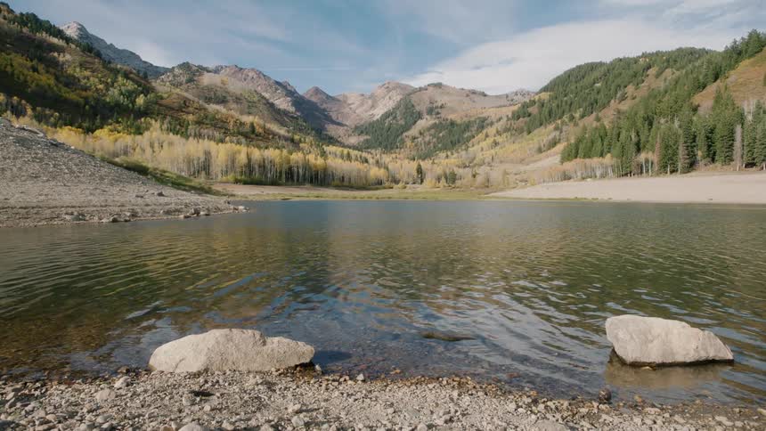 A mountain lake is surrounded by autumnal trees with yellow and green foliage set against rugged rocky mountains under a partly cloudy sky