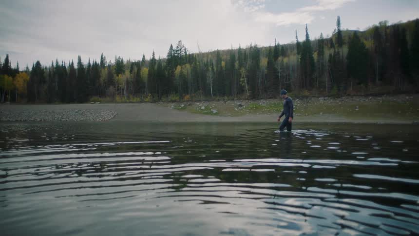 A man wearing a wetsuit wades through the shallow end of a lake on an autumn day