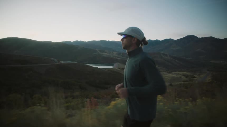 A man is jogging along a grassy rural valley with a mountain range behind it