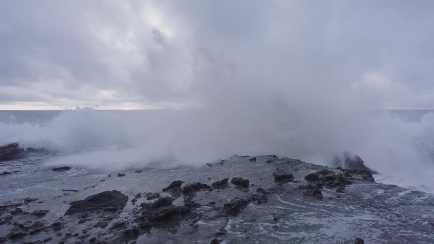 A large wave crashes on the rocks of the shore