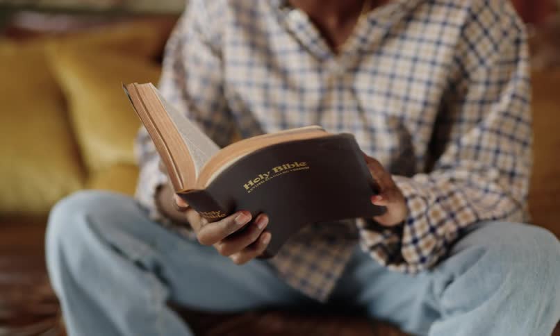 A person in a plaid shirt and jeans is sitting comfortably with a holy bible open in their hands