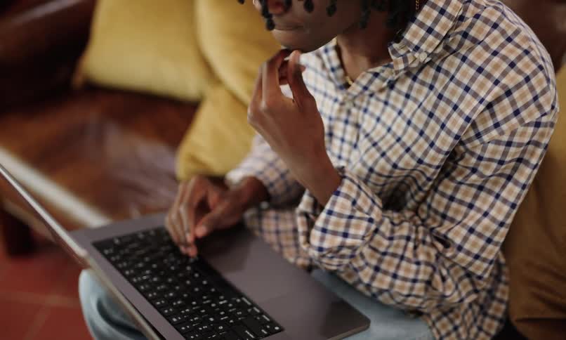 A man in a checkered shirt has his hand and chin thinking and then starts typing on a laptop that is on his lap