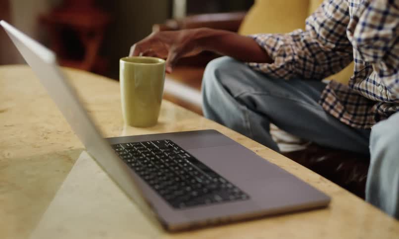 A laptop on a table and a person is fiddling with it and then he picks up a yellow mug that is next to it