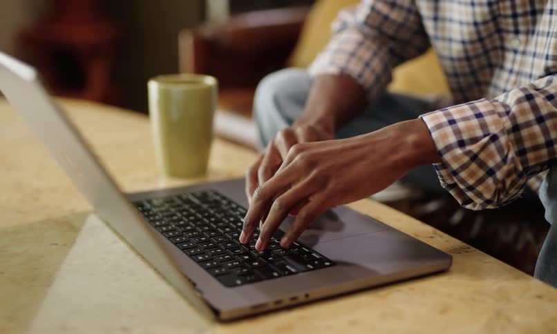 Man with his hands on a mug and then starts typing on an open laptop next to it