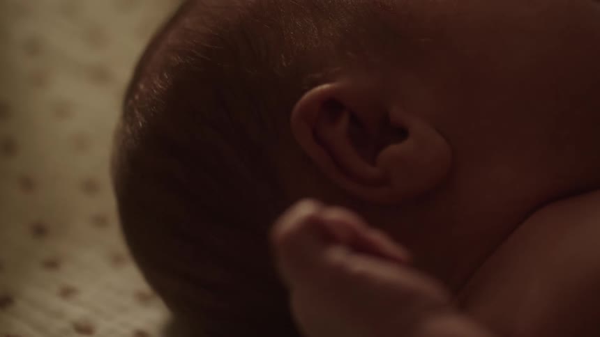 A close up shows a babys ear and clenched fist against a patterned sheet in soft lighting