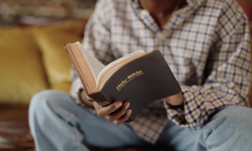 A man wearing a plaid shirt and jeans sits reading the holy bible