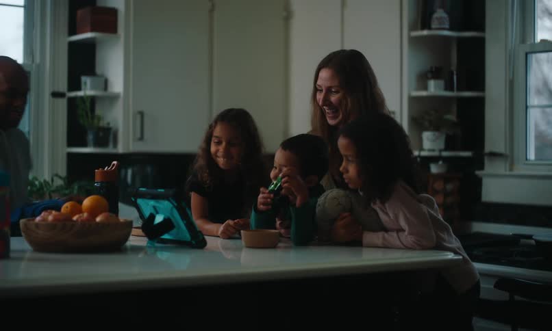 Two adults and three children are looking at a tablet on a kitchen table and smiling at it