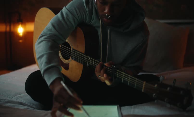 Man sitting on bed playing guitar and interacting with a tablet in front of him