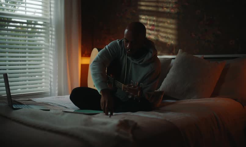 A man sits on his bed composing a song on his tablet while playing guitar in his bedroom