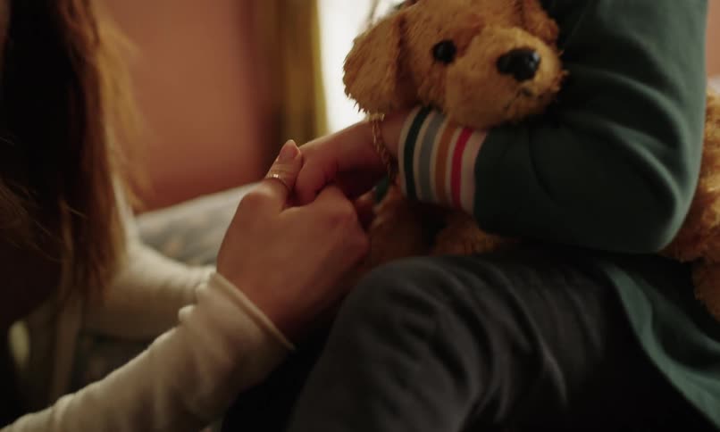 A woman with rings on her fingers is holding the hand of a child who has a stuffed dog on their lap