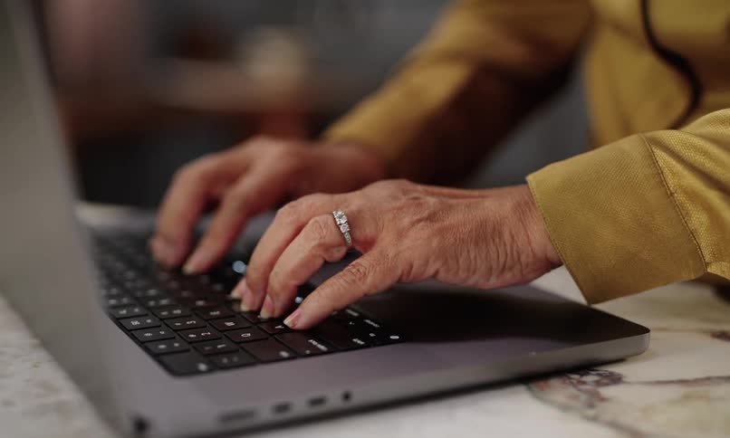 A persons hands type on a laptop keyboard with a yellow longsleeve shirt and a ring on the left hand