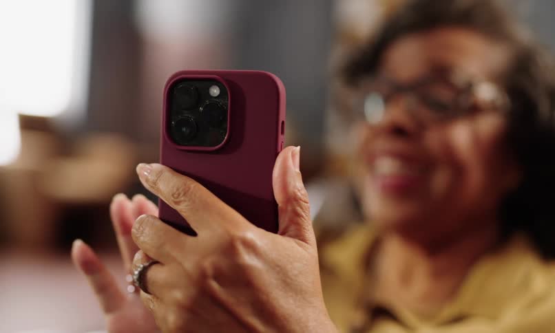 An older woman wearing glasses and a yellow shirt appears to be having a video call on her maroon cased smartphone as she gestures excitedly