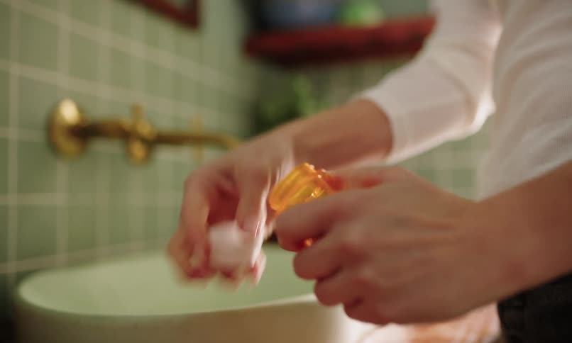 Close up of a persons hands picking up a capsule and medicine next to the bathroom sink
