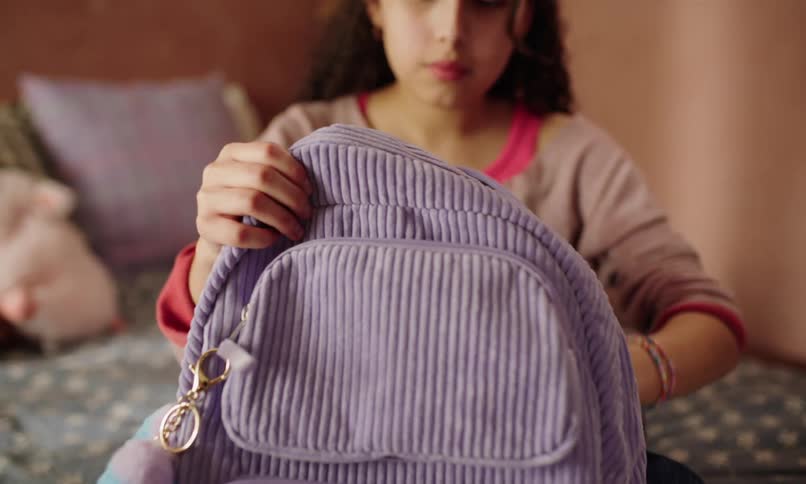 Little girl sitting on bed putting her school supplies inside her backpack