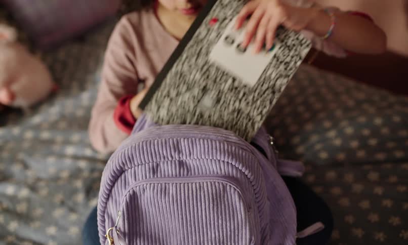 A girl with curly hair puts notebooks into her blue bag while sitting on her bed in her room