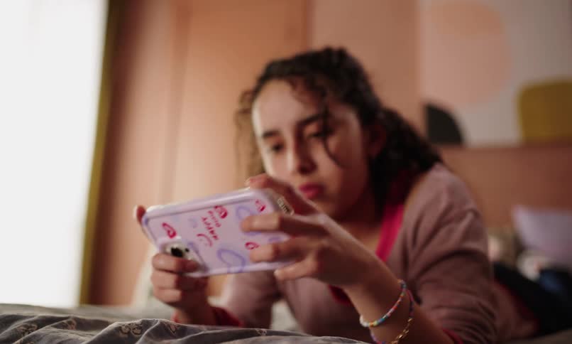 A teenage girl is lying on a bed holding and looking at a smartphone with a case that has red hearts and text on it