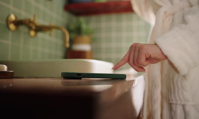 A person in a white bathrobe touches a smartphone on a bathroom counter with a brass faucet and green tiled wall in the background