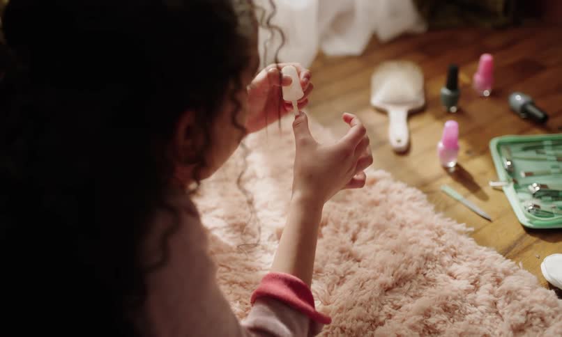 A girl with curly hair sitting on a plush rug carefully paints her fingernails surrounded by nail polish and manicure tools