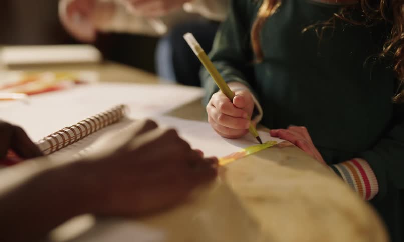 Little girl accompanied by an adult hand drawing in a notebook on the table