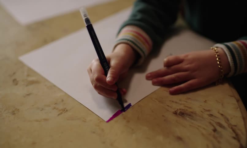 A childs hand using a purple pen to draw on a white paper