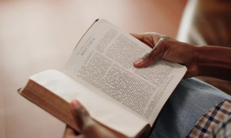 A person is holding and reading an open bible while sitting down in a room