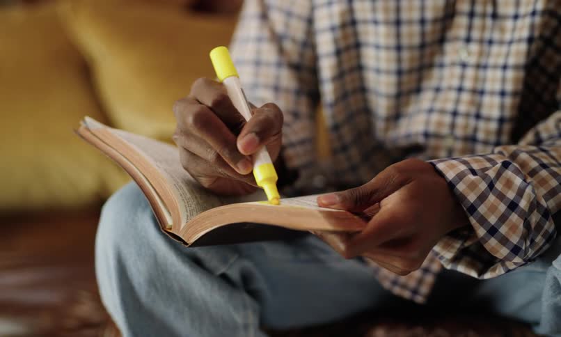 A persons hand using a yellow highlighter to mark a book they are holding