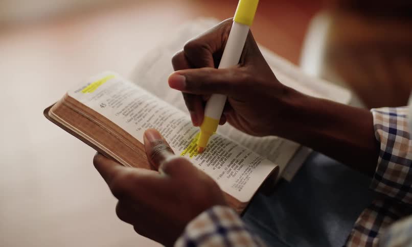 A man highlights a passage in an open book with a yellow marker their hand and a portion of their plaid patterned sleeve visible as they focus on the text