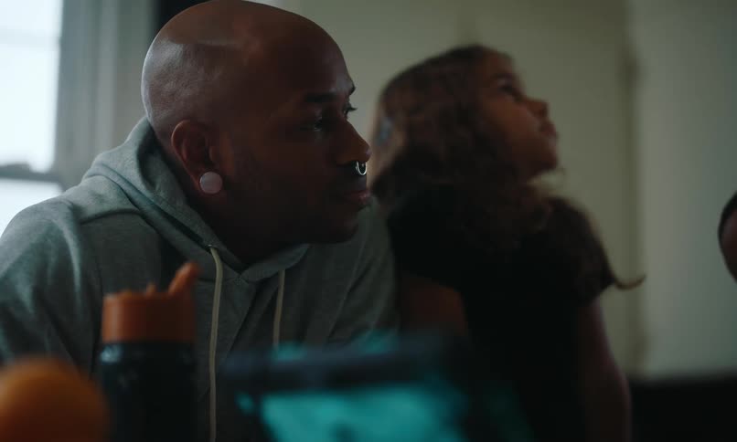 A bald man with a nose ring and a large earring in his lobe wearing a blue hoodie is sitting at a table with a child