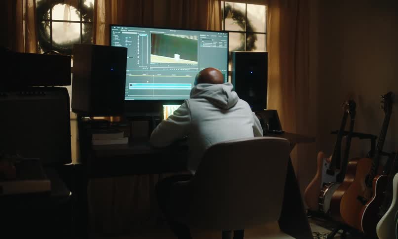 A person at a desk working on a computer with video editing software surrounded by musical instruments