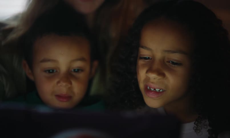 A couple of children sit on their mothers lap their faces illuminated by the screen of a laptop or tablet while watching it