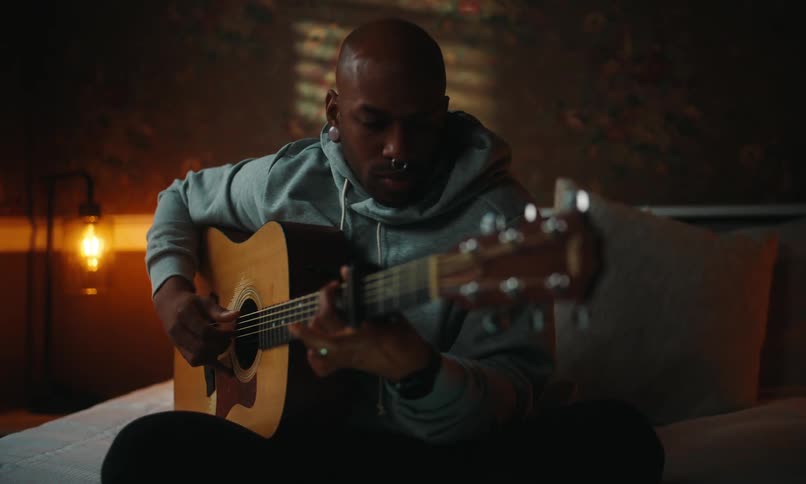 A man in a dark bedroom wearing a hoodie uses a tablet then plays music on acoustic guitar