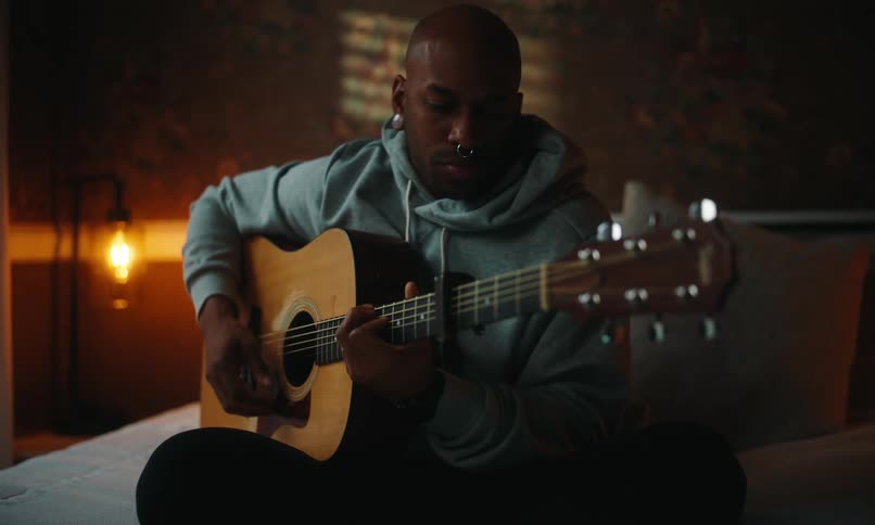 A person is sitting on a bed playing an acoustic guitar while using a stylus to interact with a tablet