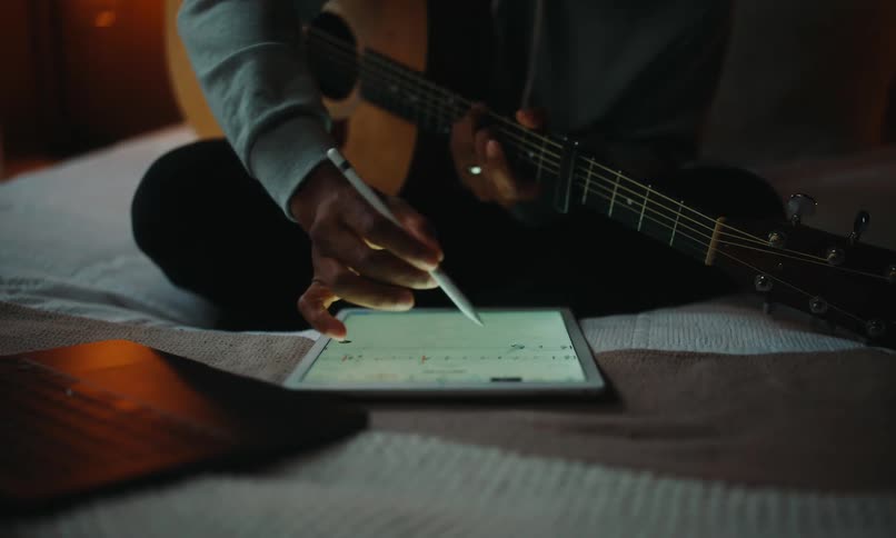 A person is sitting on a bed playing an acoustic guitar while using a stylus to interact with a tablet