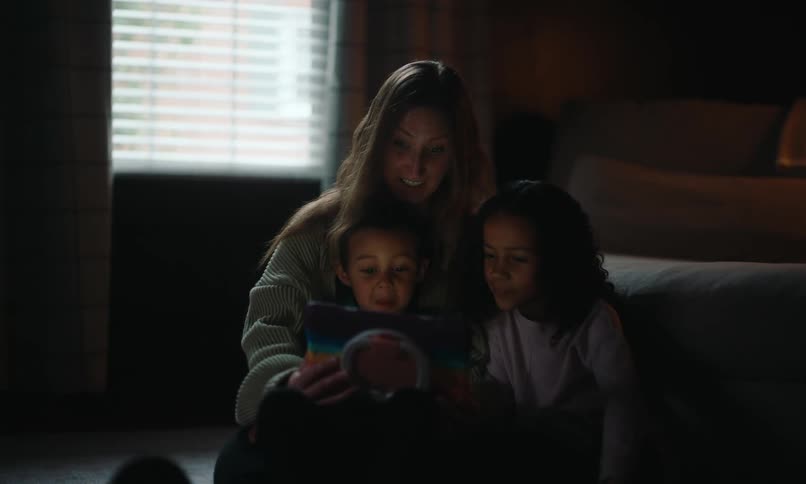 A mother and two children are sitting on the floor illuminated by the light of a tablet they are all looking at in a dimly lit room