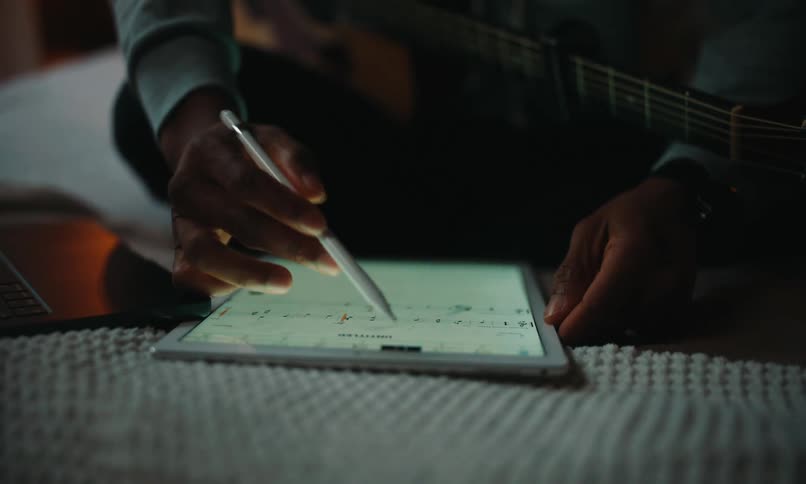 A musician sits with an acoustic guitar and uses a stylus to compose music notation on a tablet