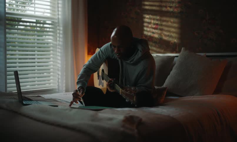 A man in a light blue hoodie composes music on a tablet while playing guitar sitting on a bed in the bedroom