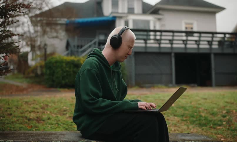 Man sitting on a bench in the backyard of the house with headphones and typing on a laptop