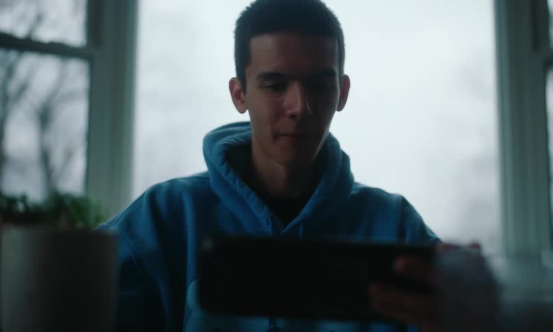 A person in a blue hoodie is drinking from a cup while holding a tablet in a dimly lit room with large windows in the background