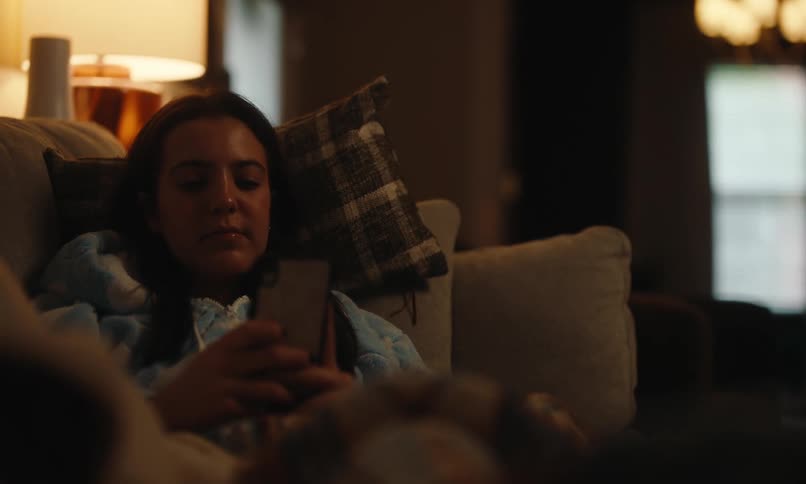 Woman relaxes on the couch snuggled up in a sweatshirt while using her cell phone in a warmly lit room