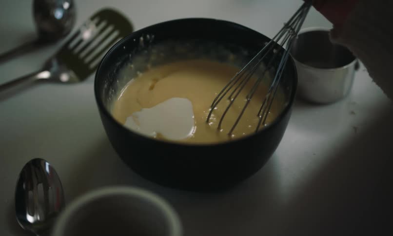 Woman pours milk from a measuring cup into a bowl of yellow batter while using a whisk