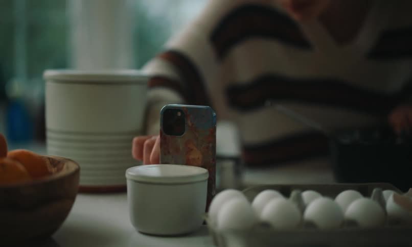A woman wearing a striped knit sweater is preparing breakfast in a kitchen while looking at a recipe on her smartphone