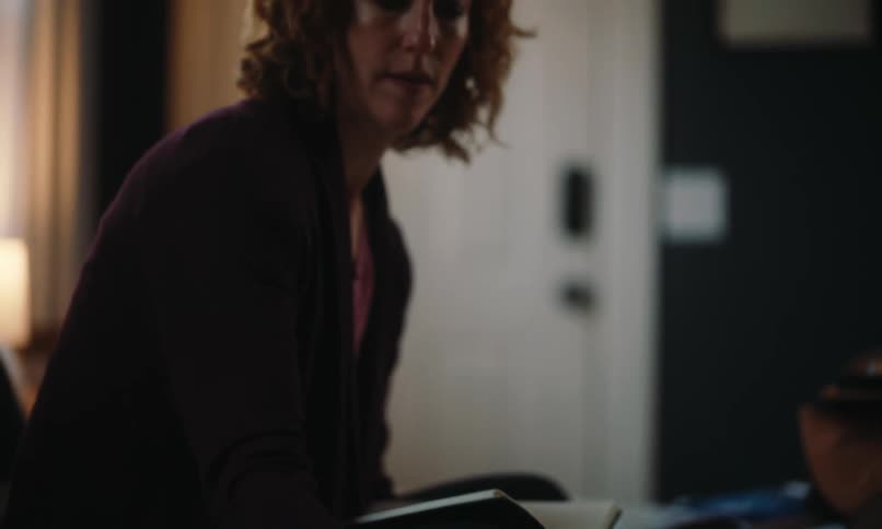 Sitting on her bed a red-headed woman with curly hair checks her iphone while working on her laptop and notebook