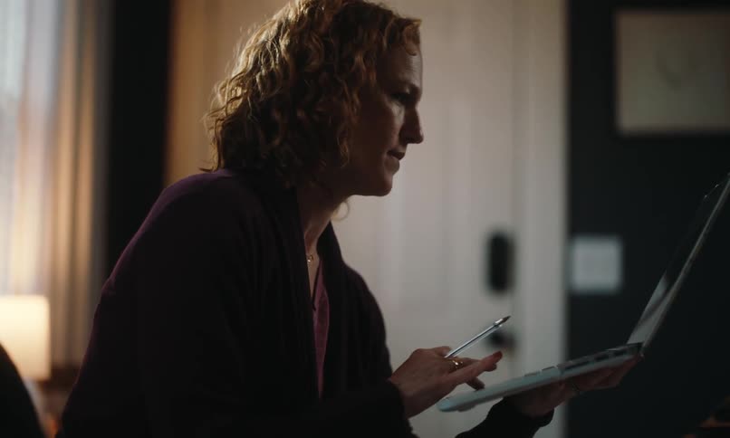 A woman with curly hair sits in a dimly lit room talking to someone and interacting with her laptop screen
