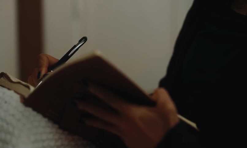 A woman with glasses wearing wireless earbuds and a black sweater writes in a notebook while a white blanket rests on her lap