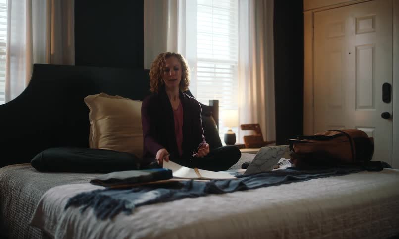 A woman sits cross legged on a bed with a laptop and journal appearing to be working or studying in a brightly lit room