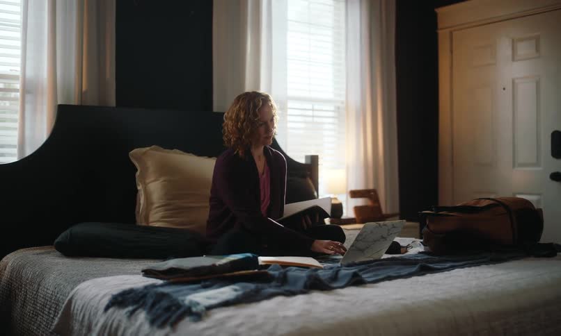 A woman is sitting crosslegged on a bed in a dimly lit room surrounded by books papers and a laptop engaged in studying or working