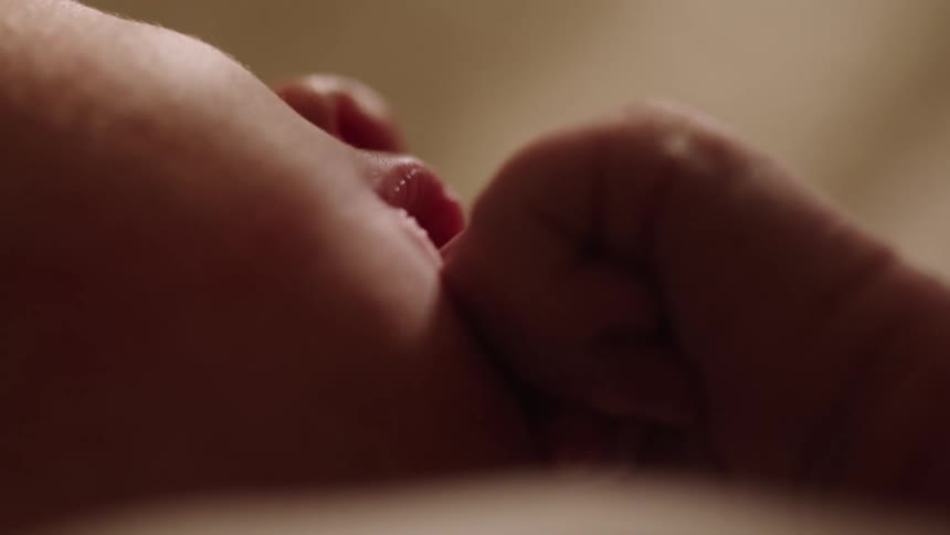 A closeup of a baby resting in a dimly lit room with their hand on their mouth